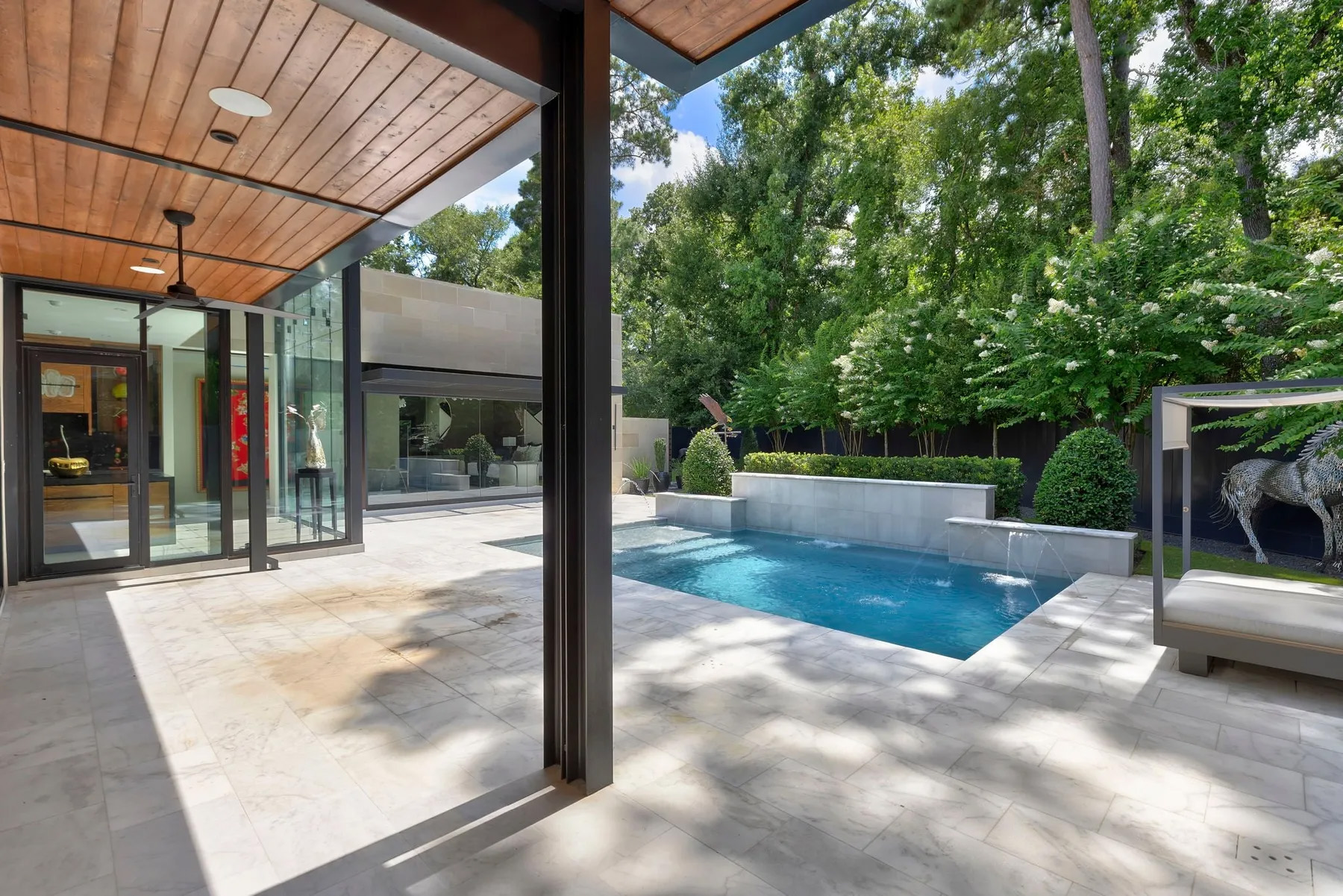 Modern backyard with a small rectangular pool, stone patio, glass walls, wood ceiling, manicured shrubs, and tall trees in the background on a sunny day—perfect for hosting an A-list gathering in August 2026.