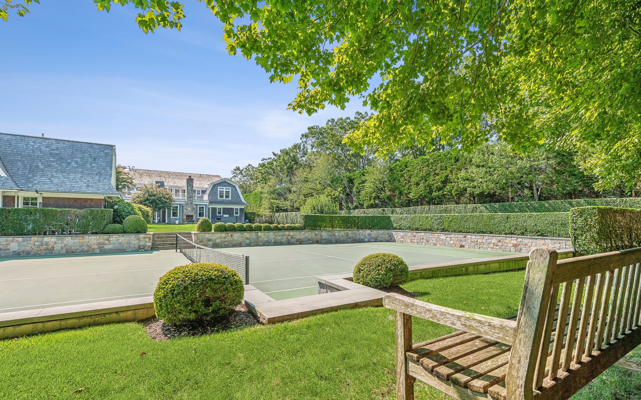 A wooden bench overlooks a well-maintained outdoor tennis court surrounded by lush greenery, manicured bushes, a stone wall, and large houses in the background under a clear blue sky—an inviting setting that perfectly showcases the summer amenities of this vibrant neighborhood.