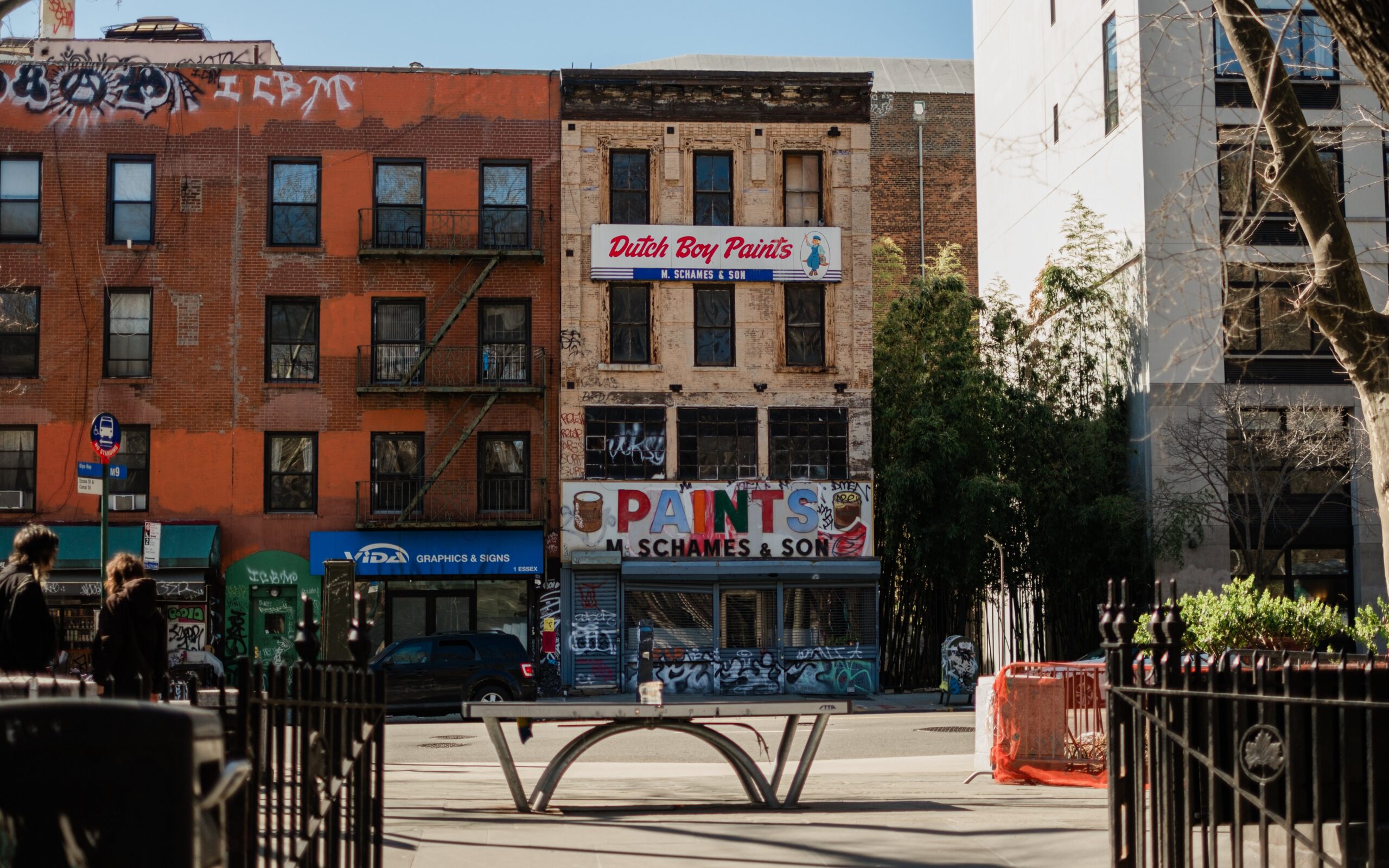 A city street on the Lower East Side with a weathered Dutch Boy Paints storefront, graffiti, apartment buildings, and a metal skateboard ramp in the foreground. People walk along the sidewalk under clear daylight.