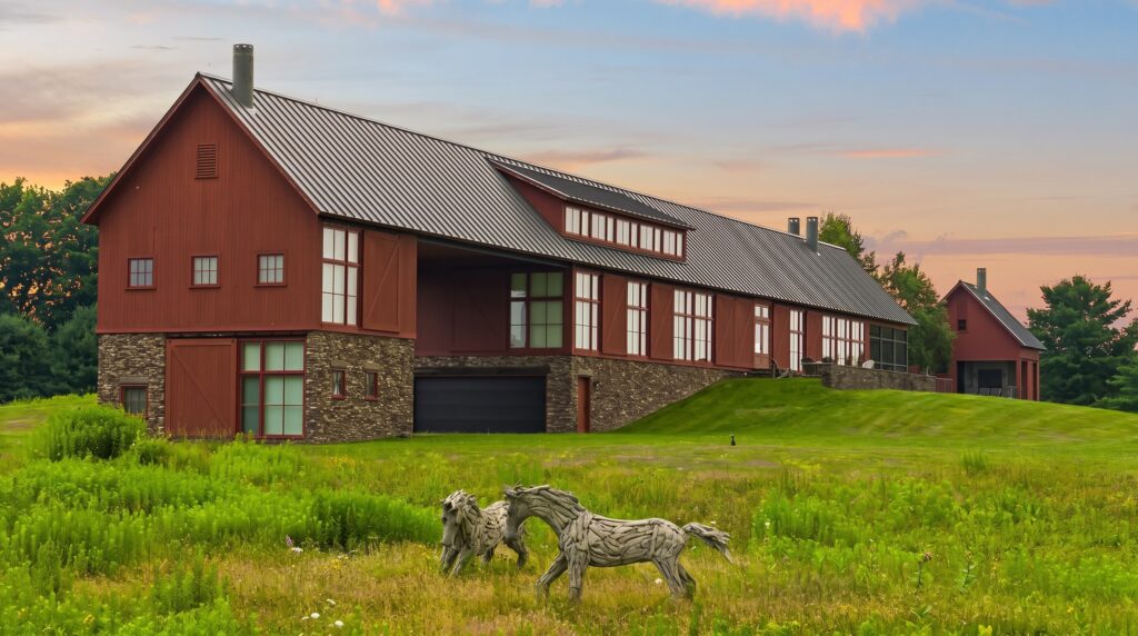 Barn-style luxury estate with a separate guest house, stone accents, expansive lawns, and sculptural horse artwork, showcasing modern farmhouse architecture in a scenic countryside setting.