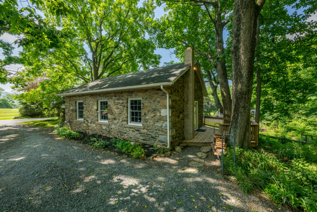 Historic stone guest house surrounded by mature shade trees and lush landscaping, featuring rustic masonry construction, a gravel driveway, and a serene woodland setting.