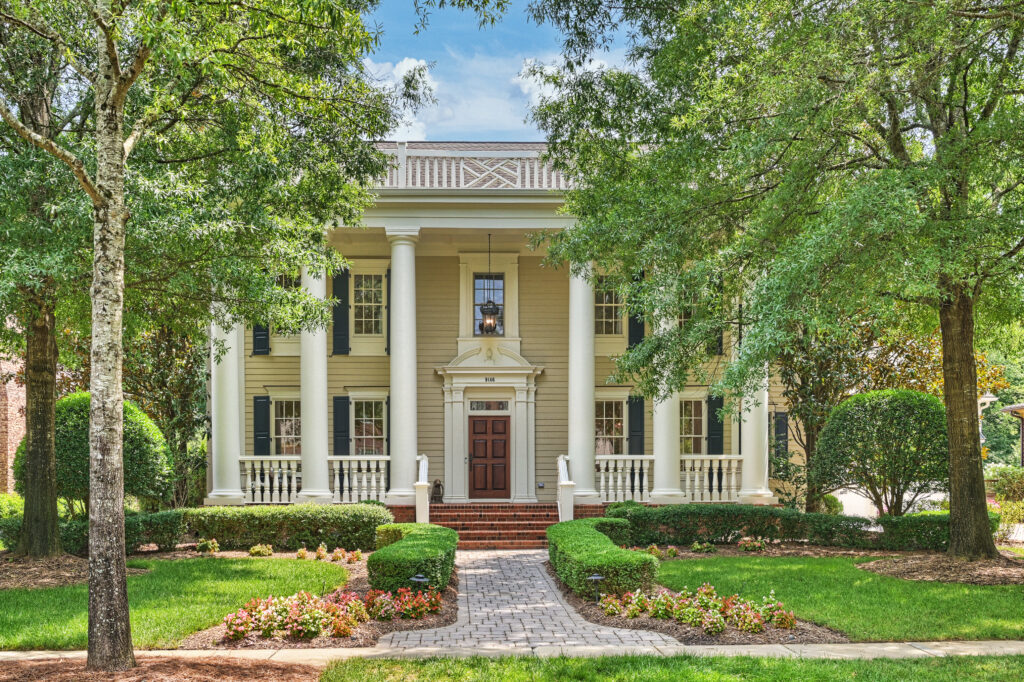 Libra A two-story beige house with white columns, a brown front door, brick steps, and manicured landscaping is framed by tall trees and a lush green lawn under a blue sky—perfectly embodying the tidy elegance often associated with Virgo season.