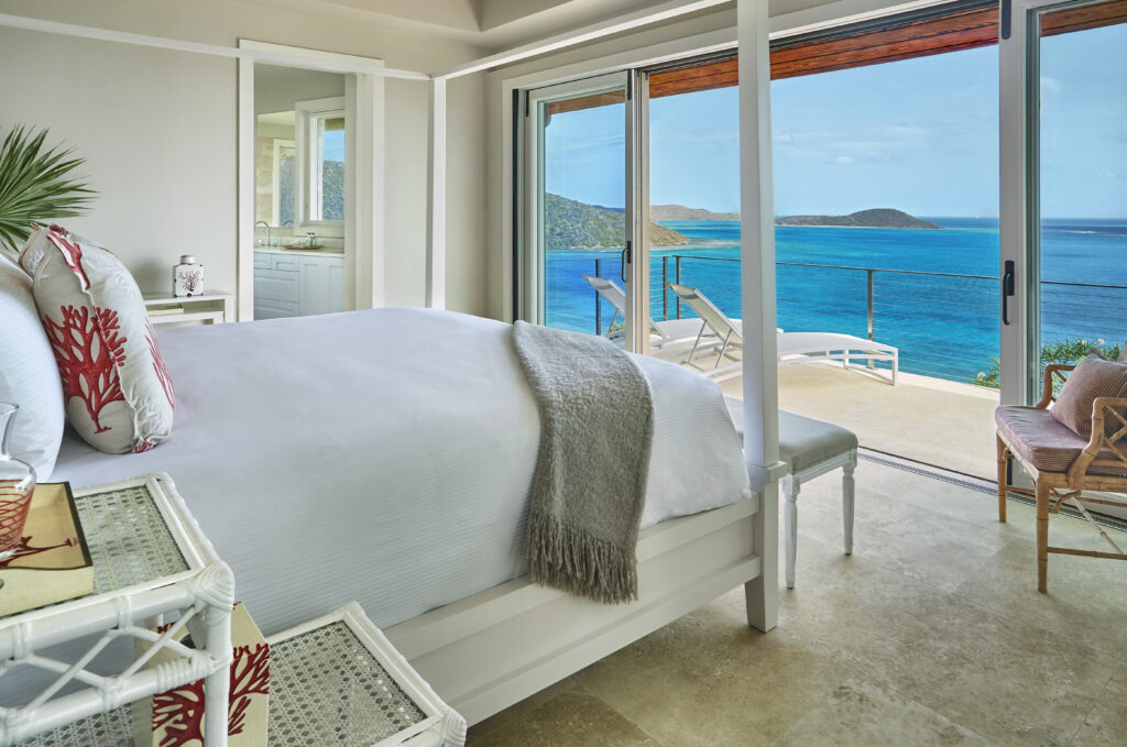 Luxury oceanfront guest house bedroom with a four-poster bed, floor-to-ceiling sliding glass doors, private terrace, and panoramic turquoise water views, showcasing upscale Caribbean-style indoor-outdoor living.