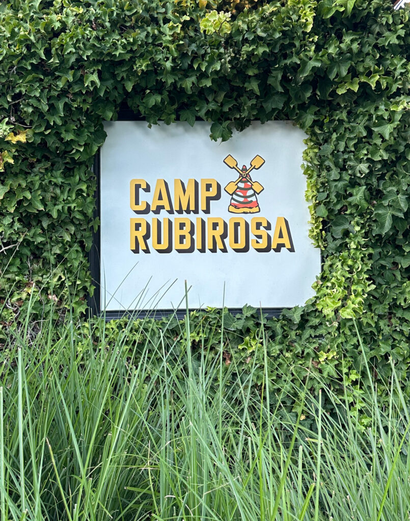 A sign reading Camp Rubirosa with a camp hat and paddles illustration, surrounded by green ivy and tall grass.