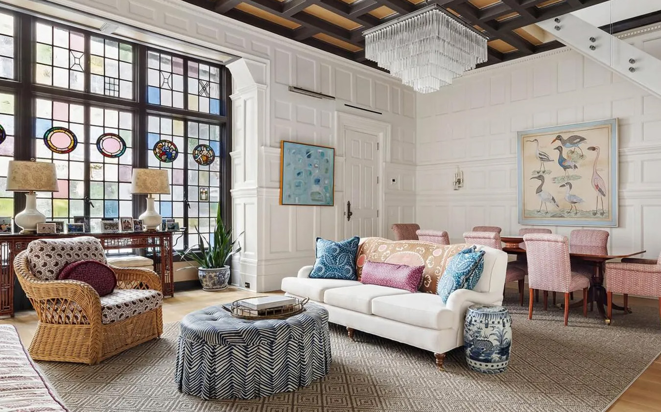 Bright living room with large stained glass windows, white paneled walls, a white sofa, patterned armchair, blue ottoman, and a dining area with pink chairs. Artwork and decorative accents add color and interest.