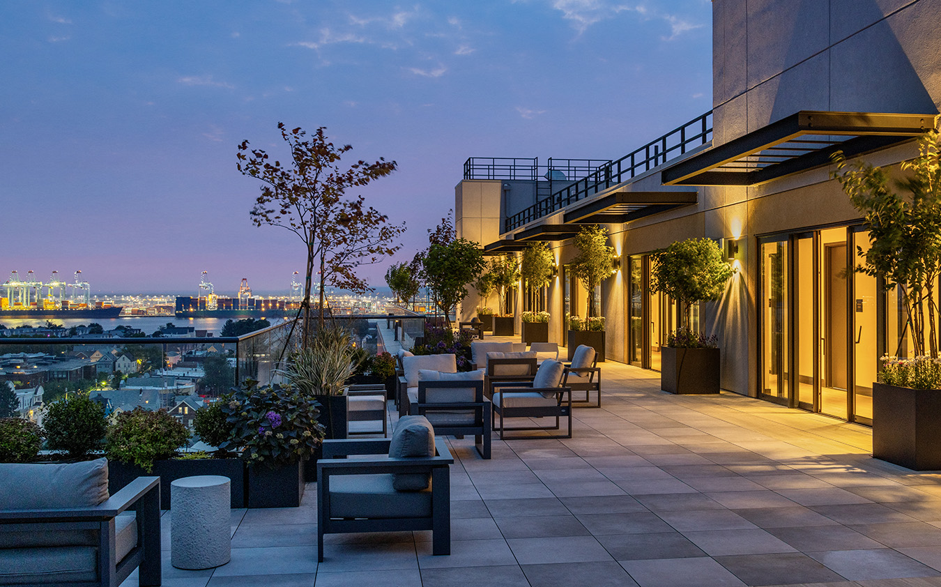 Modern rooftop terrace at dusk with outdoor seating, potted plants, glass railing, and cityscape views. Warm lights glow from inside the building under a partly cloudy sky.