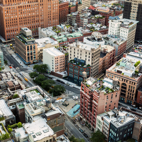 Aerial view of a city block with mid-rise buildings, tree-lined streets, intersections, and cars. Rooftops show various structures and greenery, characteristic of a nyc residential rental neighborhood. The scene has a mix of modern and older architecture.