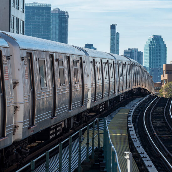 A silver subway train curves along elevated tracks in a city, passing by tall buildings, skyscrapers, and the occasional townhouse, all visible in the background under a clear blue sky.