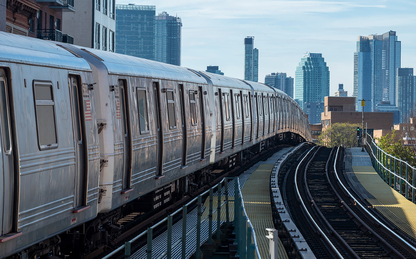 A silver subway train curves along elevated tracks in a city, passing by tall buildings, skyscrapers, and the occasional townhouse, all visible in the background under a clear blue sky.