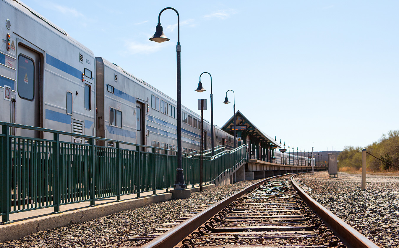 A silver passenger train is stopped at a small outdoor station with a green fence, lampposts, and a covered platform beside the train tracks under a clear blue sky.