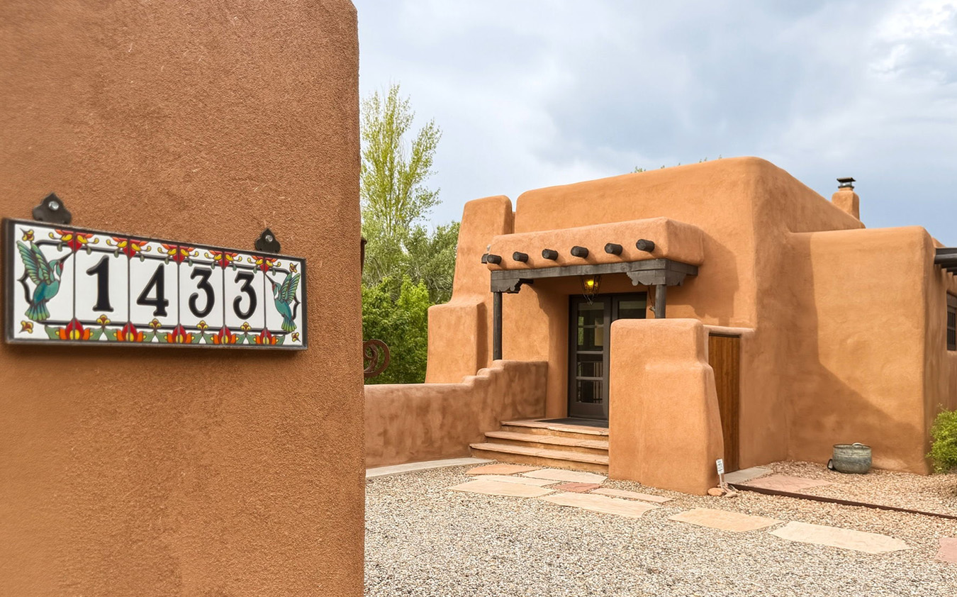 th_santafe A tan adobe-style Santa Fe house with wooden beams and steps leading to the front door. In the foreground is a tile address plaque with the number 1433. The sky is cloudy and trees are visible in the background.