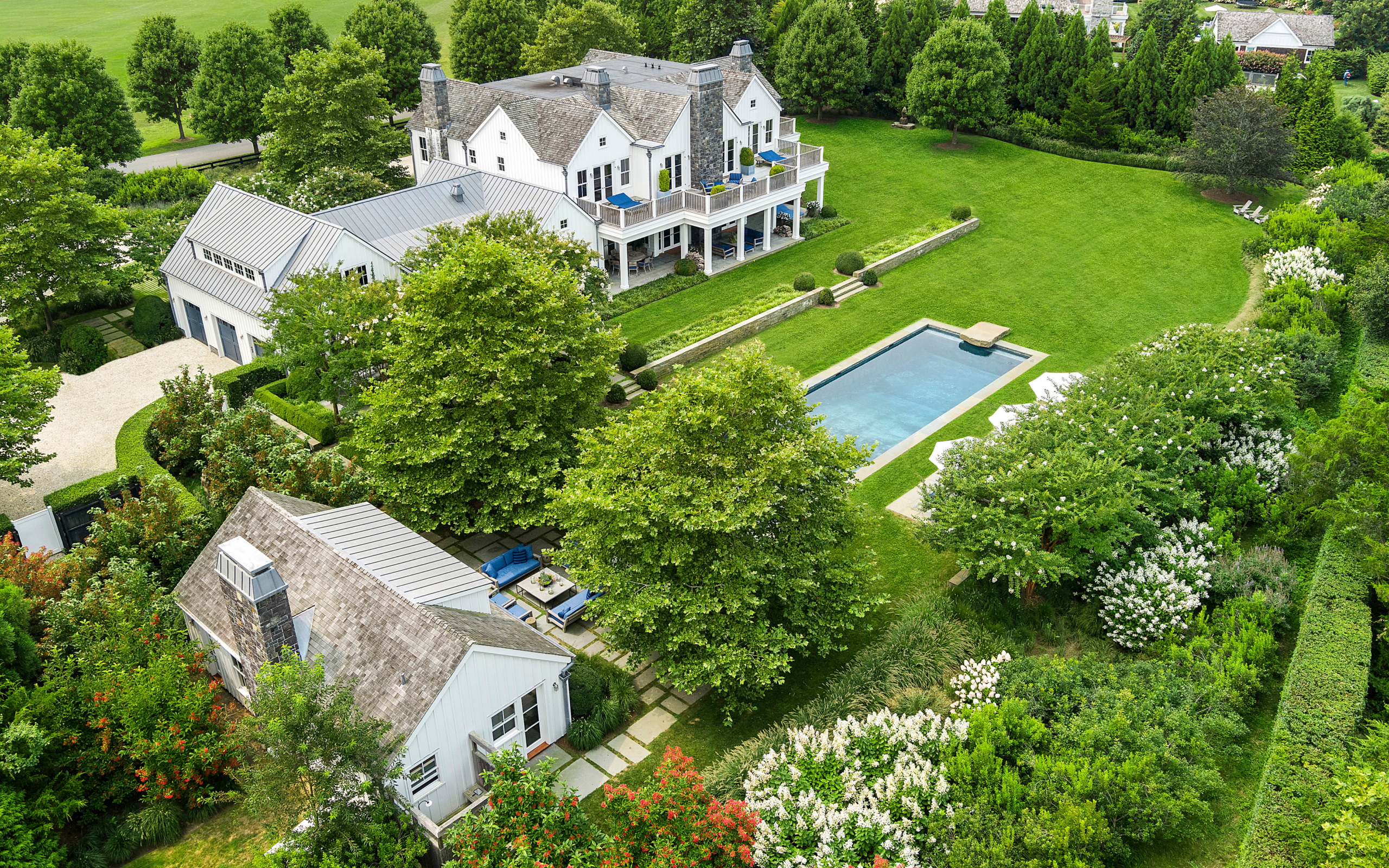 Aerial view of a large white house with balconies, a guest house, a rectangular pool, green lawns, trees, and landscaped gardens on the spacious 12 Two Trees Lane property.