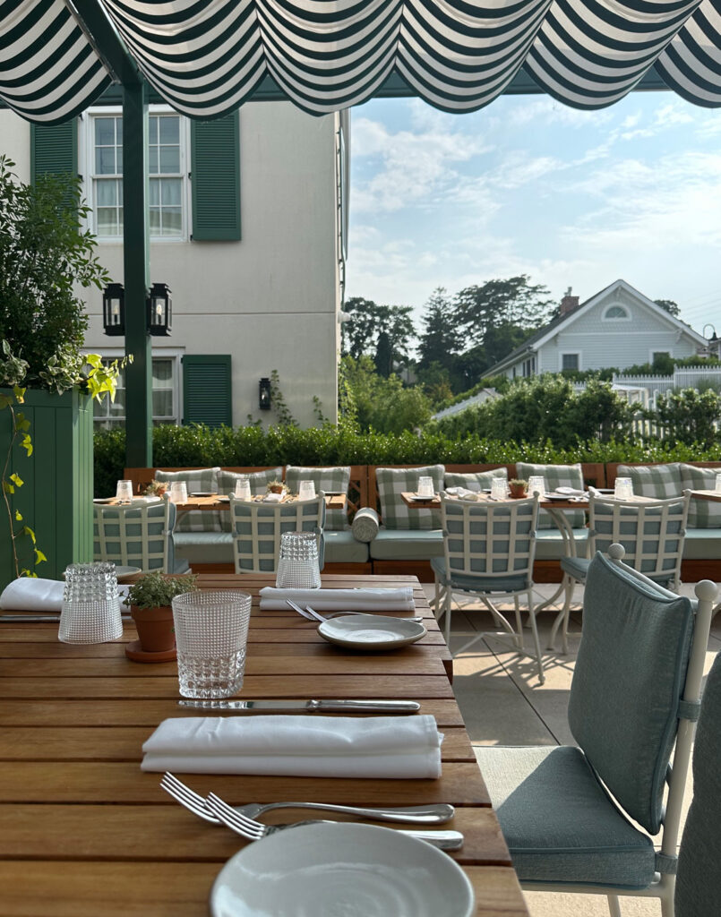 A sunlit outdoor dining area with a wooden table set for four, green-striped chairs, potted plants, and a striped canopy. In the background are white buildings with green shutters and lush greenery.