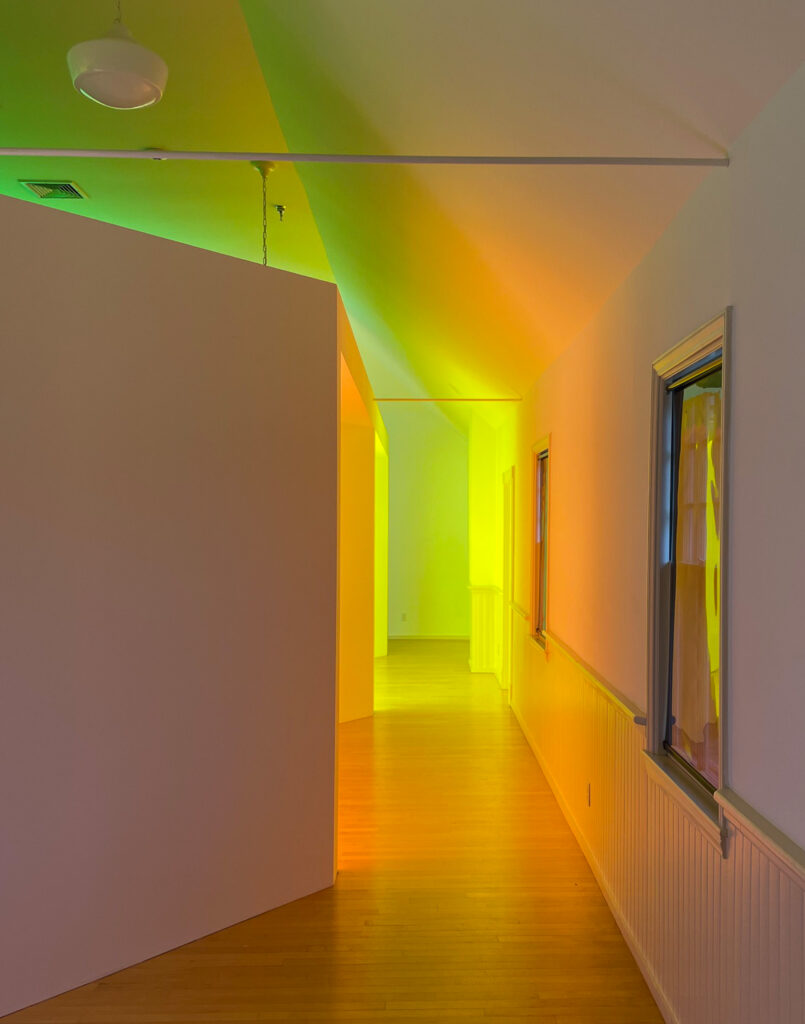 A hallway with wooden floors and white walls is illuminated by yellow, orange, and green lights, creating a gradient effect on the walls and ceiling.