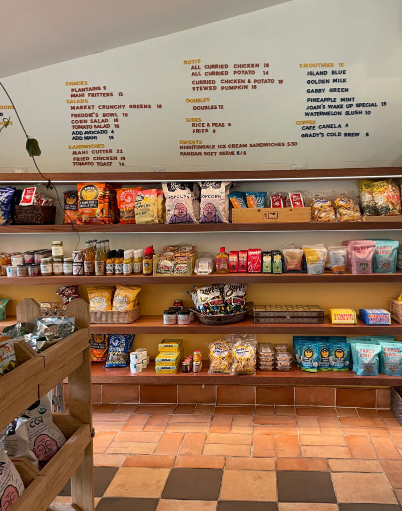 Small grocery store with shelves of snacks, chips, and packaged goods below a menu board listing food and drink options. Earth-toned tiles on the floor and natural light brighten the scene.