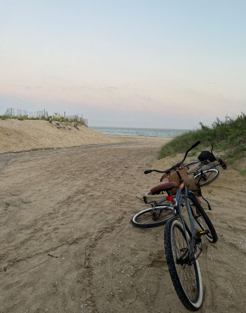 Two bicycles rest on a sandy path leading to the beach, bordered by grassy dunes, with the ocean visible in the distance under a soft, pastel sky.