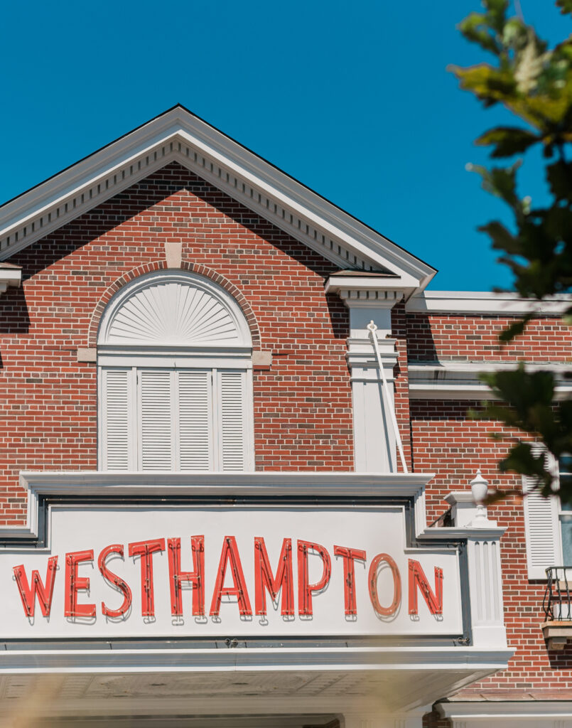A bright red Westhampton sign is mounted on a brick building with white trim and closed shutters, set against a clear blue sky.