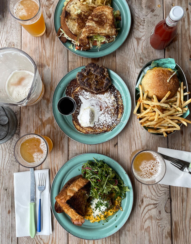 Three plates with breakfast foods—scrambled eggs with toast, pancakes with sausage, and a burger with fries—are set on a wooden table, accompanied by drinks and utensils.