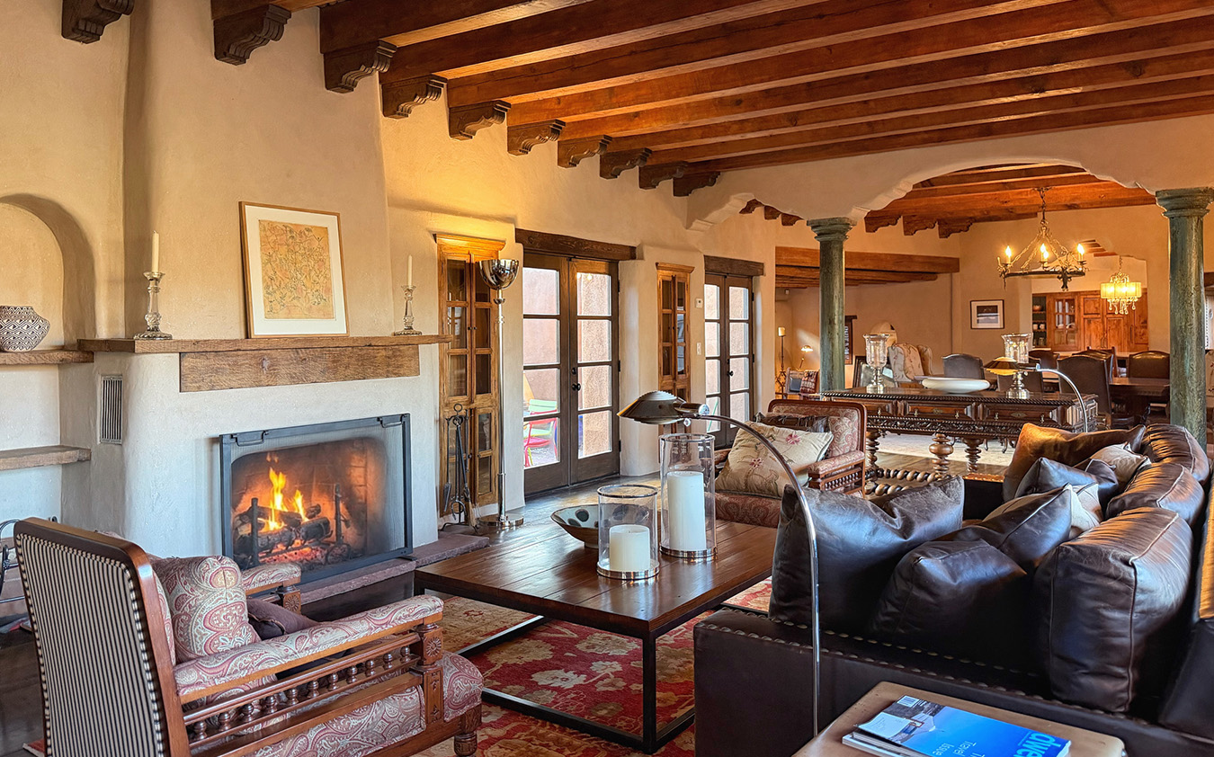 A cozy living room in a classic Santa Fe house with exposed wooden beams, a lit fireplace, leather sofas, armchairs, a large coffee table, and French doors. Warm lighting and rustic decor create an inviting, comfortable atmosphere.