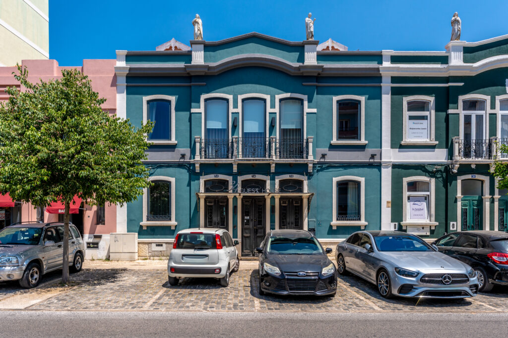 Three cars are parked in front of a blue two-story building with arched windows and statues on the roof, under a clear blue sky—a setting that perfectly captures the balanced energy of libra season. A leafy tree stands on the left side of the image.
