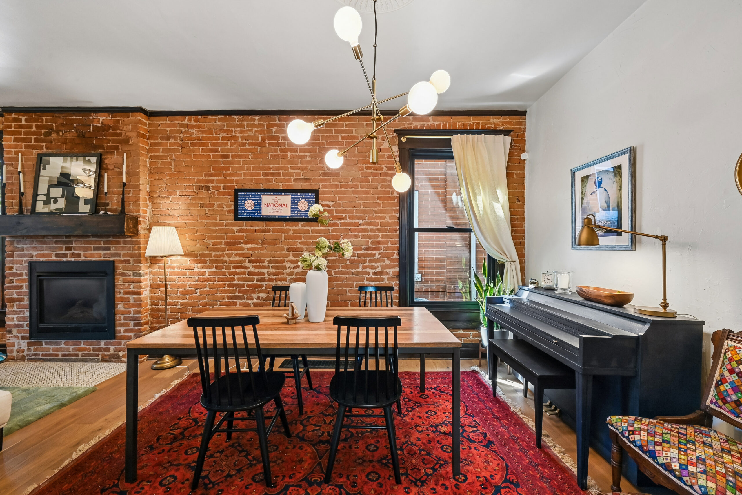 Dining room with exposed brick walls, a black piano, modern chandelier, black chairs, a wooden table with vases, a red rug, a fireplace, artwork, and a window with white curtains letting in natural light—a space designed for refined gatherings worthy of an A-list: September 2026 soirée.