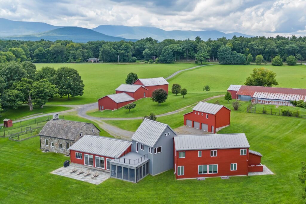 Aerial view of a modern farmhouse complex with red and gray buildings, green lawns, scattered trees, and distant mountains under a partly cloudy sky—a tranquil retreat that feels perfectly balanced, as if designed in harmony with Libra season.