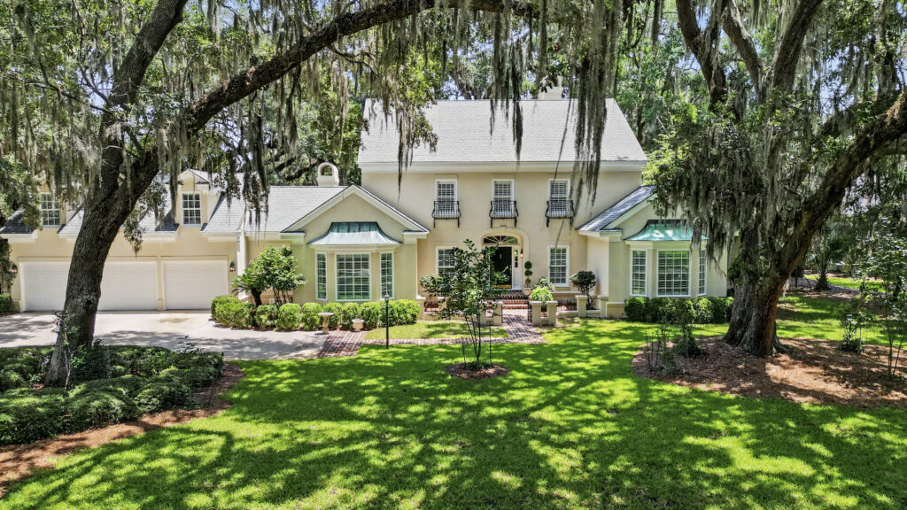 Large cream-colored house with three garages, tall windows, and French-style balconies, framed by large moss-covered trees casting shadows on a manicured green lawn—a serene retreat perfectly suited for the harmony of Libra season and the elegance often attributed to astrological houses.
