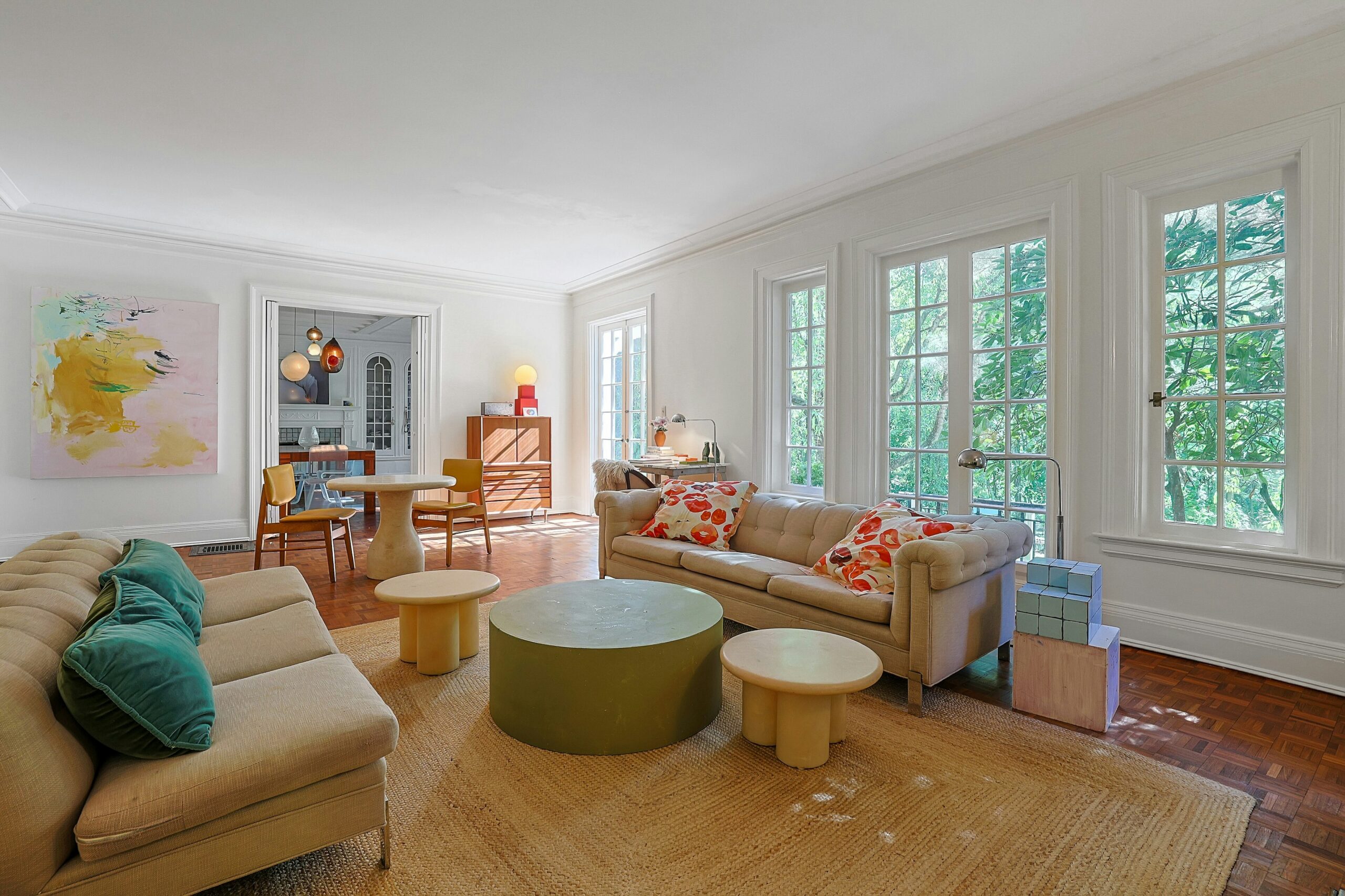Bright living room with large windows, beige sofas with colorful pillows, a round green coffee table, wooden floor, and abstract wall art—an inviting space perfect for hosting an A-list gathering in September 2026. The dining area in the background features modern lighting for a chic finishing touch.