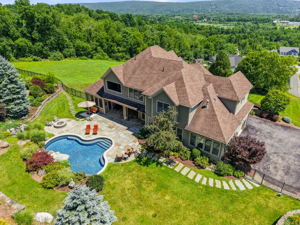 A large house with a tan roof sits amid greenery, featuring a landscaped backyard, patio with chairs, pool, hot tub, and curved stone path—an inviting scene that feels especially harmonious during Libra season, with hills and trees in the background inspiring balance and tranquility.