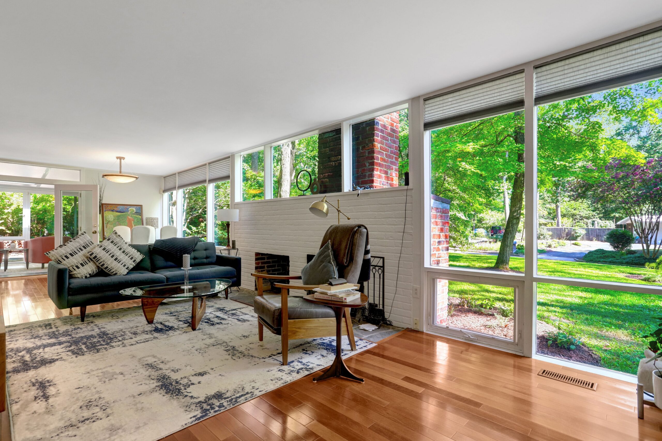 Mid-century modern living room with large windows, black sofa, glass coffee table, armchair, brick fireplace, wood floors, and a view of green trees outside—an a-list style space that feels ready for September 2026.