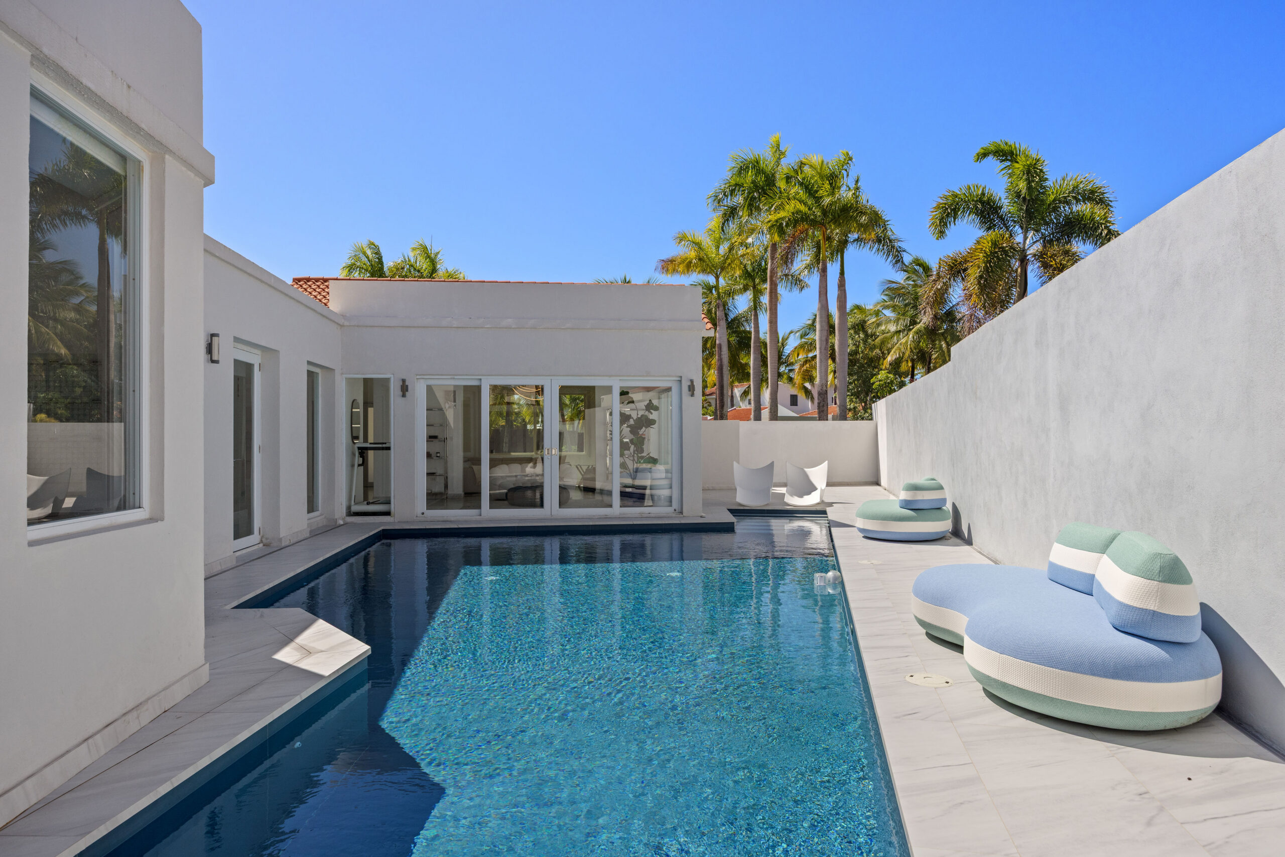 Modern backyard with a rectangular pool, blue and white lounge chairs, white walls, large windows, palm trees, and clear blue sky—an inviting retreat designed for relaxing or entertaining like an a-list celebrity. Experience resort-style living perfect for hosting friends and family in September 2026 and beyond.