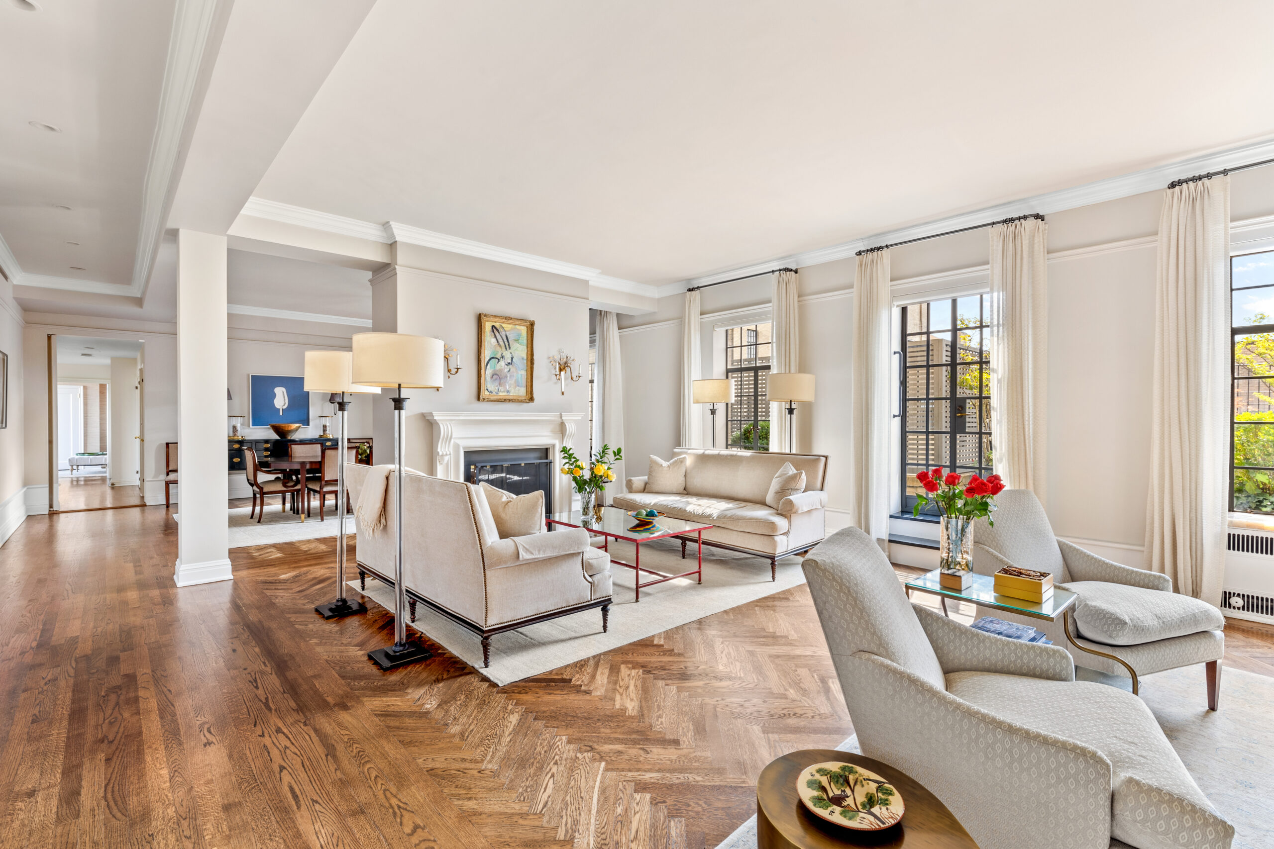 Spacious, sunlit living room with wood floors, white sofas, armchairs, a fireplace, large windows with curtains, and fresh flowers on tables—perfect for stylish entertaining or relaxing evenings. With its elegant design and inviting atmosphere, this A-list retreat is ready to welcome you in September 2026. Dining area visible in the background.