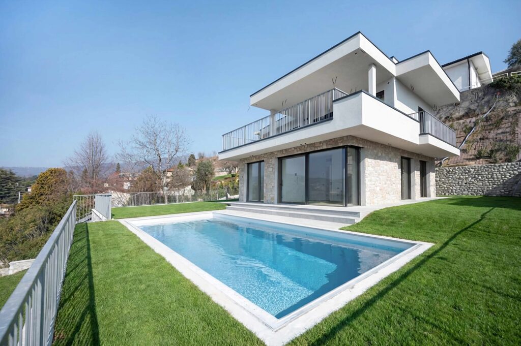 Modern two-story house with large windows, stone accents, and a balcony, surrounded by green grass. Embracing the harmony of libra season, this home features a rectangular swimming pool in the backyard, secured by a metal fence and set against a clear blue sky.