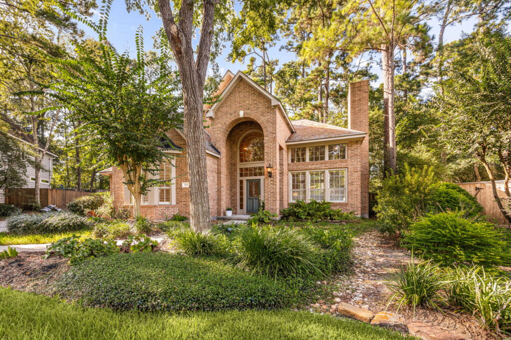 Two-story brick house with arched entryway, green front yard, large trees, and landscaping. Tall windows and a chimney are visible, surrounded by lush greenery and a wooded background—an inviting retreat that feels especially harmonious during Libra season.