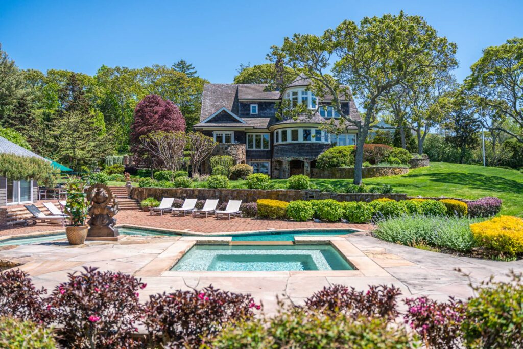 Large house with multiple gables and a turret, surrounded by trees and manicured gardens. In the foreground, there is a hot tub and swimming pool area with lounge chairs on a brick patio—an inviting retreat that feels especially harmonious during Libra season, when balance and beauty are celebrated in astrological houses.