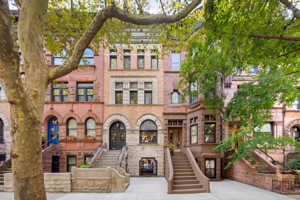 A row of historic brownstone townhouses with ornate facades, tall windows, and stairs leading to the entrances, shaded by leafy green trees on a quiet city street—each residence as beautifully balanced as an astrological house during Libra season.