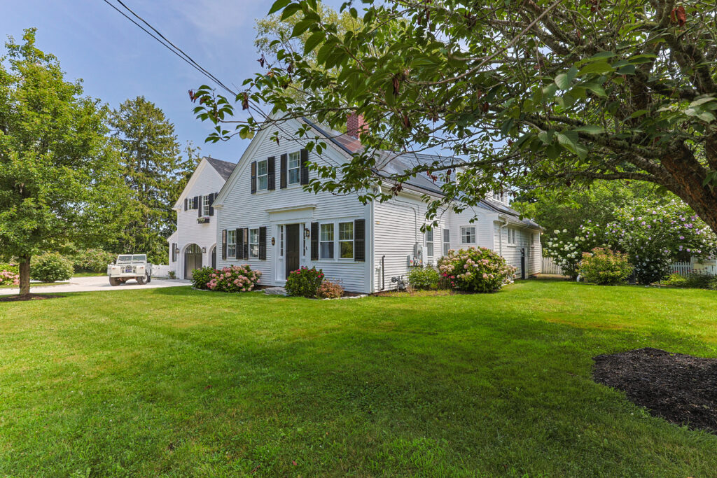 White two-story house with dark shutters and attached garage, surrounded by green lawn, trees, and blooming bushes on a sunny day—a welcoming retreat that feels especially harmonious during Libra season, inviting balance and beauty to every corner.