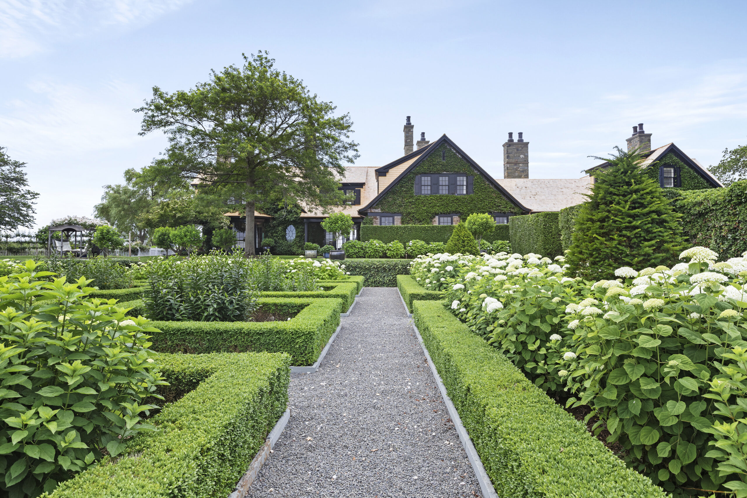 A gravel path leads through neatly trimmed hedges and blooming white hydrangeas toward a large house with a dark exterior, surrounded by lush greenery under a clear blue sky—a picture-perfect retreat worthy of any A-list gathering in September 2026.