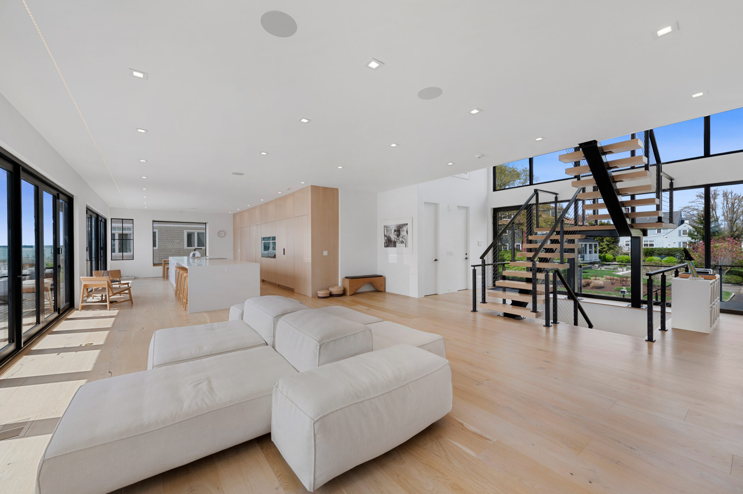 Spacious modern living area with light wood floors, white furniture, open kitchen, large windows, and a floating staircase leading upstairs. Natural light fills the room, and there’s a view of a landscaped yard outside—an ideal setting for an A-list gathering in September 2026.
