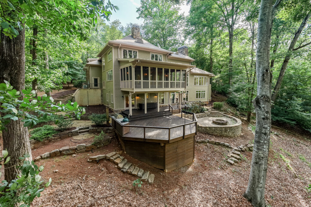 Large beige house with multiple stories, screened porch, and wooden deck set in a wooded area. The backyard features a circular stone fire pit with curved stone seating amid tall trees and natural landscaping—an ideal retreat for stargazing or reflecting on the astrological houses during peaceful Libra season.