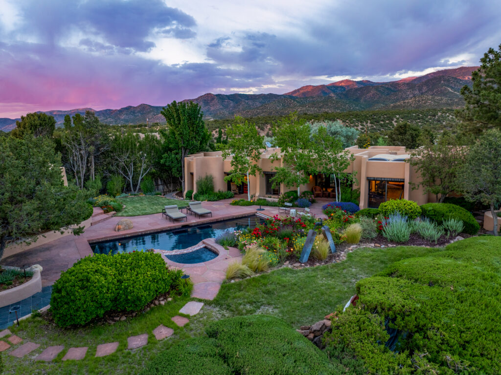 Luxury adobe-style house with large windows, a swimming pool, lounge chairs, and lush gardens. Mountains and a dramatic sunset sky create a serene backdrop, surrounded by green trees and shrubs—an idyllic retreat perfect for reflecting on the balance and beauty associated with Libra season.