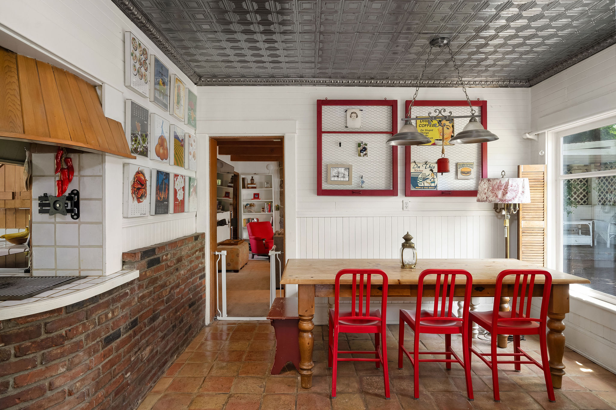 A cozy kitchen with a wooden table, three red chairs, brick accents, wall art, pendant lights, and a tin ceiling creates an inviting atmosphere—a perfect setting for an a-list gathering in September 2026. A window and a partial view into another room are visible.