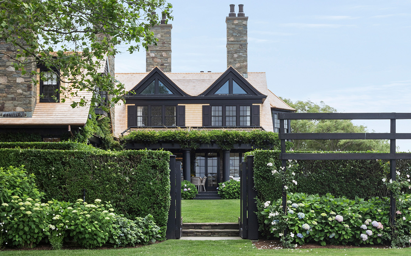 A large stone house with black trim, two chimneys, and manicured hedges. There’s a gated entrance, lush green lawn, and flowering bushes in the foreground beneath a partly cloudy sky—perfectly capturing sophisticated curb appeal worthy of an A-list estate: September 2026.