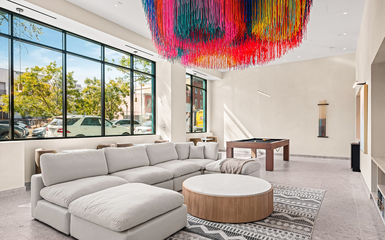 Modern lounge with a large white sectional sofa, round wooden coffee table, pool table, and a vibrant multicolored hanging art fixture beneath tall windows with street and tree views.