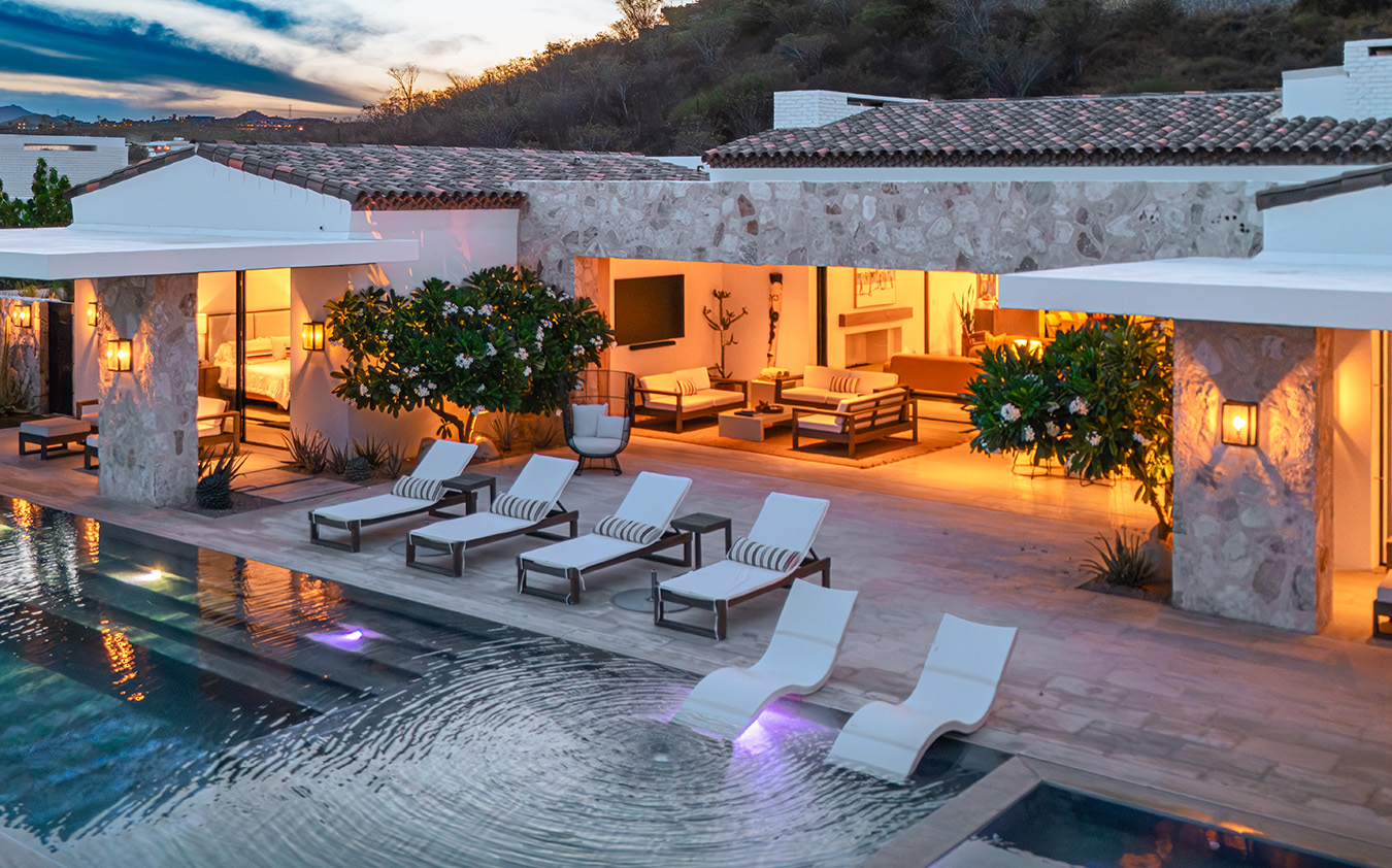Modern villa with poolside lounge chairs, illuminated patio, and outdoor seating area at sunset; stone walls, lush plants, and tranquil water create a luxurious, relaxing atmosphere.