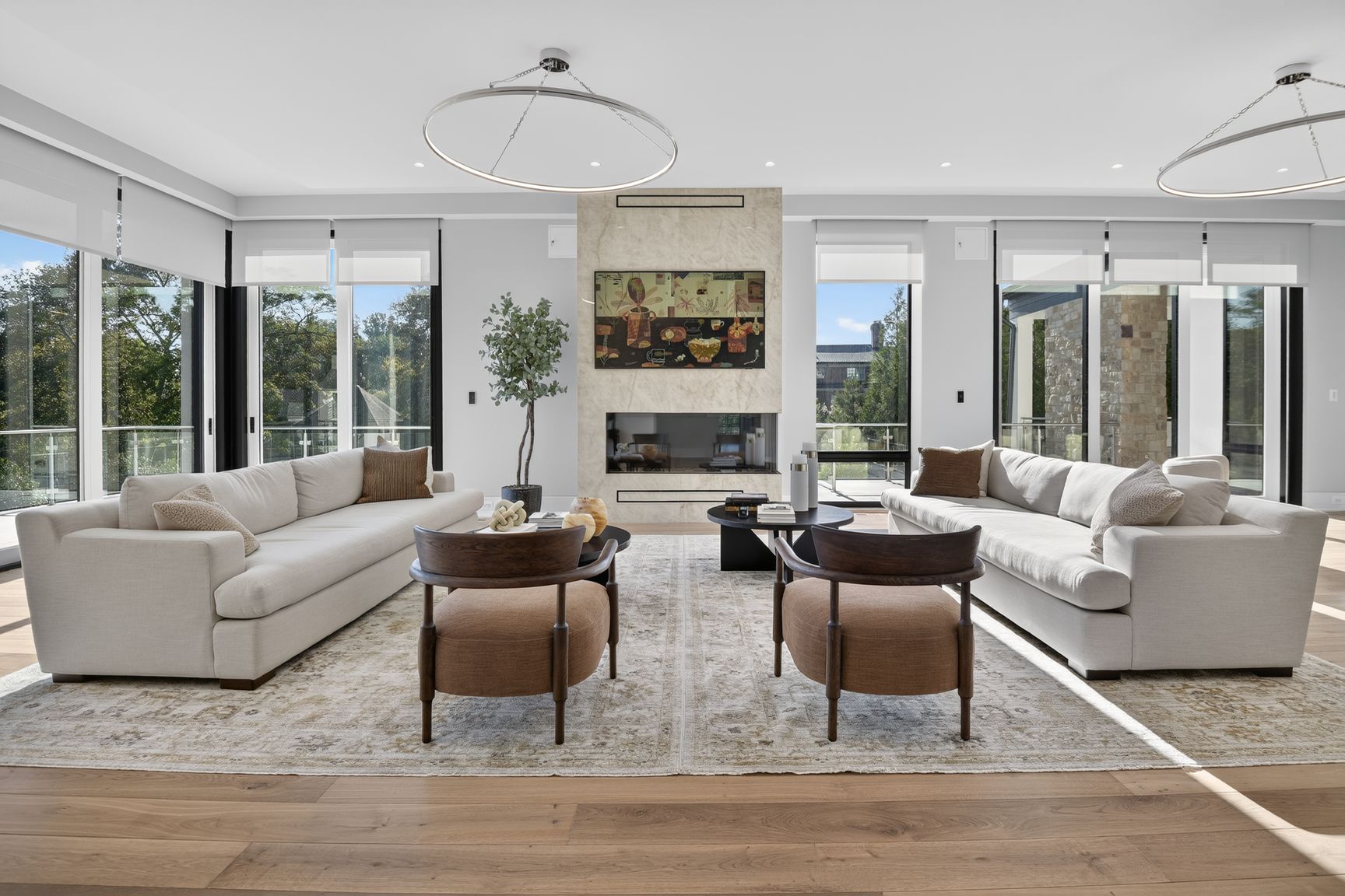 A bright, open-plan living room featuring two facing beige sofas, two round armchairs, a central fireplace with mounted artwork, and floor-to-ceiling windows, as seen in the A-List November 2026.