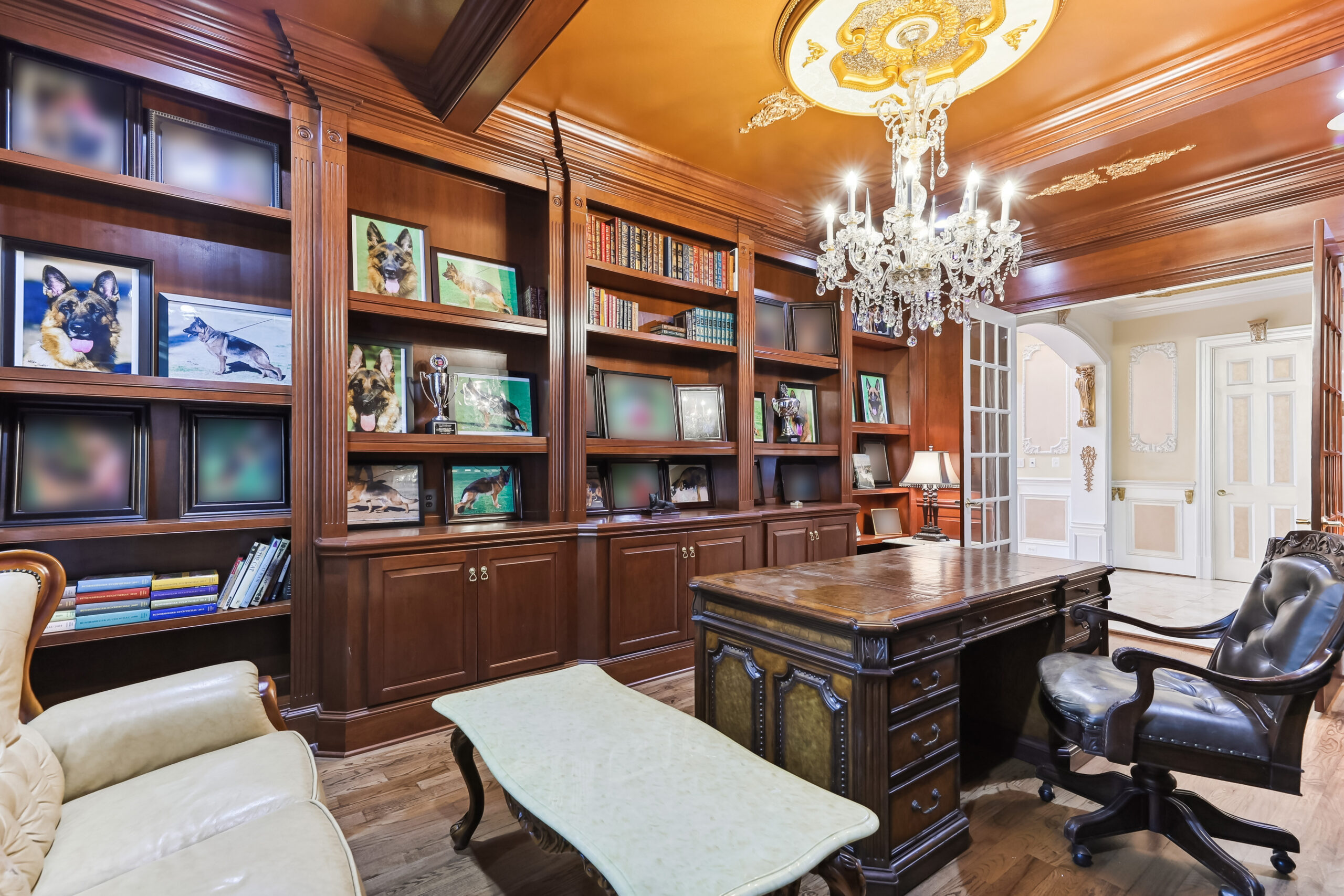 12792 Yates Ford Rd A home office with dark wood built-in bookshelves displaying framed dog portraits and trophies, featuring a large wooden desk, a leather office chair, and a crystal chandelier.