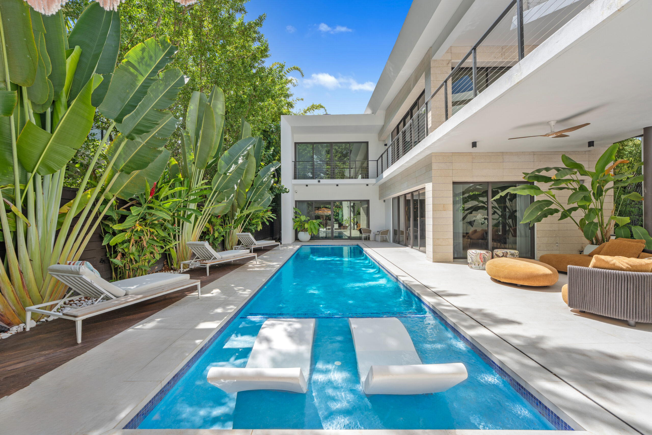 A rectangular swimming pool with two submerged lounge chairs is surrounded by a modern two-story home, poolside chairs, and lush tropical plants, perfect for an October 2026 a-list getaway.