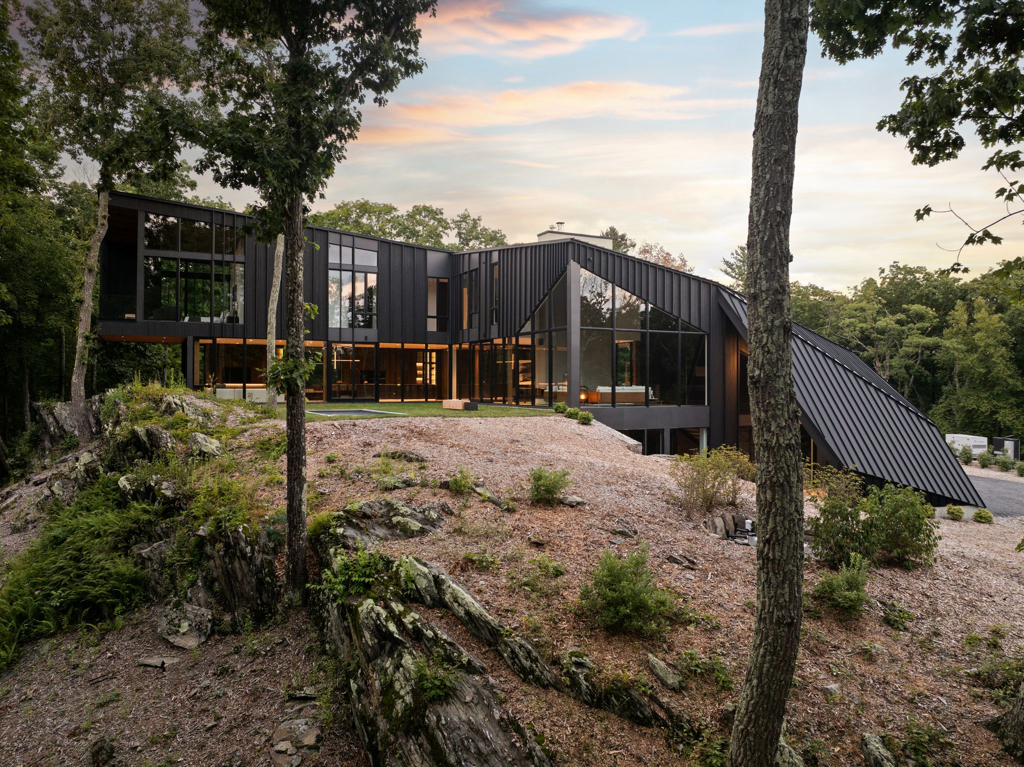 A modern, dark-sided multi-level home with large windows built into a rocky, wooded hillside at sunset, captured in October 2026.