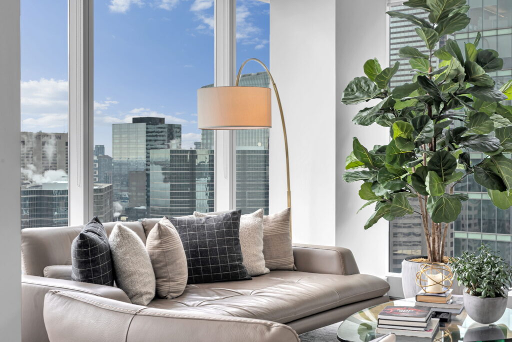 183 Wellington Street W 3303 Toronto C01, ON M5V-0A1 Toronto C01, ON M5V-0A1 A leather sofa with patterned throw pillows sits in a guest house in front of a floor-to-ceiling window overlooking a city skyline, with an arched floor lamp and a large potted plant nearby.